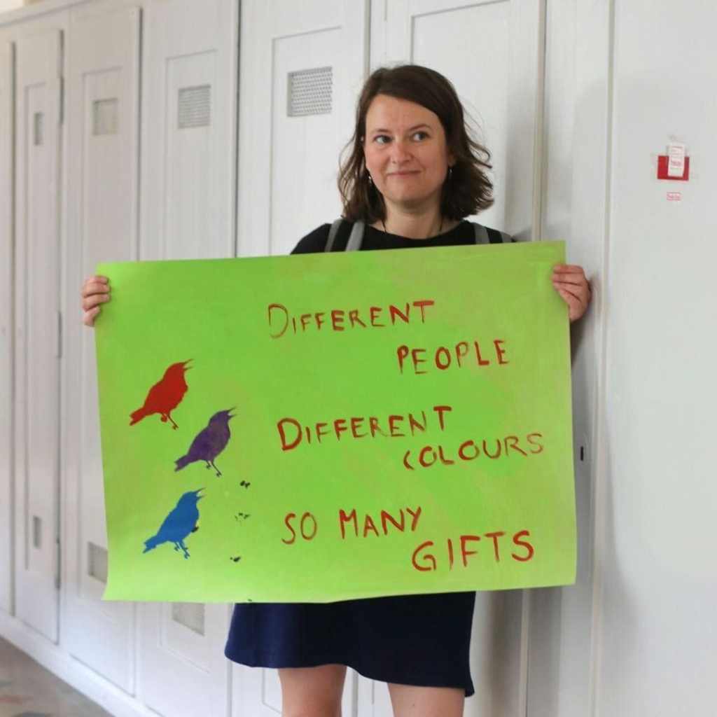 Ich halte ein hellgrünes Plakat in den Händen. Darauf steht von Hand geschrieben: "Different people, different colours, so many gifts"

Links vom Text sind 3 Vögel in 3 Farben gedruckt.